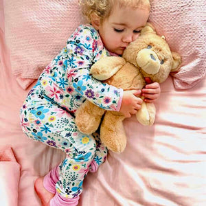 Child in floral pajamas hugging a teddy bear on a pink blanket