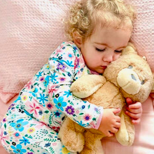 Child in floral pajamas holding a teddy bear on a pink blanket