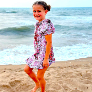 Young girl in a floral dress standing on a sandy beach with ocean waves in the background.