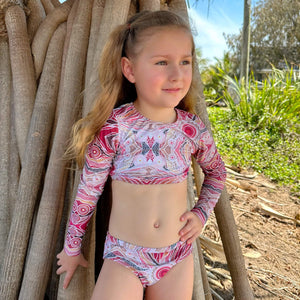 Young girl in a colorful swimsuit standing next to a woven structure with greenery in the background
