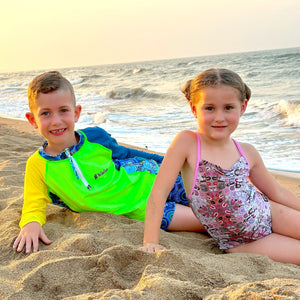Two children, a boy and a girl, sitting on a sandy beach with ocean waves in the background.