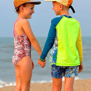 Two children standing on a log by the ocean, wearing swimsuits and hats.