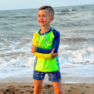 Child in bright swim shirt and shorts standing on a beach with ocean waves in the background